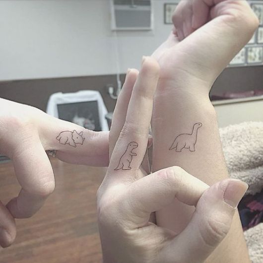 Tatuagens de dinossauros entre os dedos e pulso de três pessoas diferentes. 