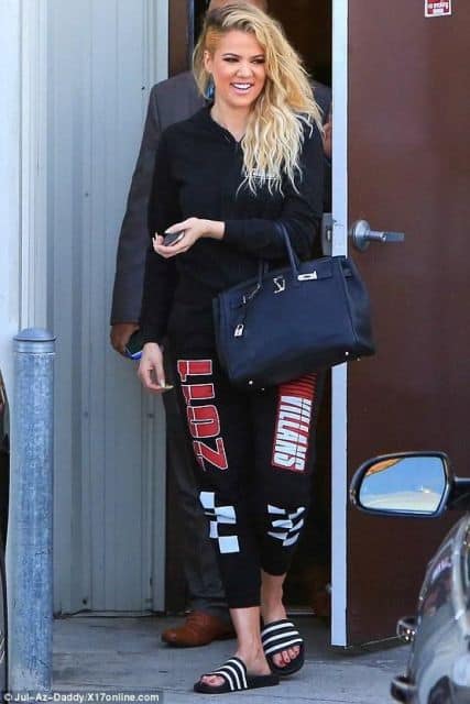Khloe Kadarshian veste calça moletom estampada, blusa preta , bolsa e chinelo slide no mesmo tom.