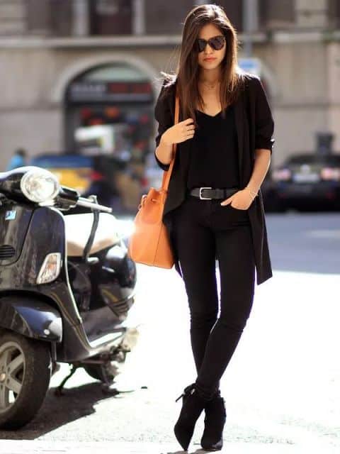 Modelo usa blusa, casaquinho, calça e bota preta com bolsa laranja.