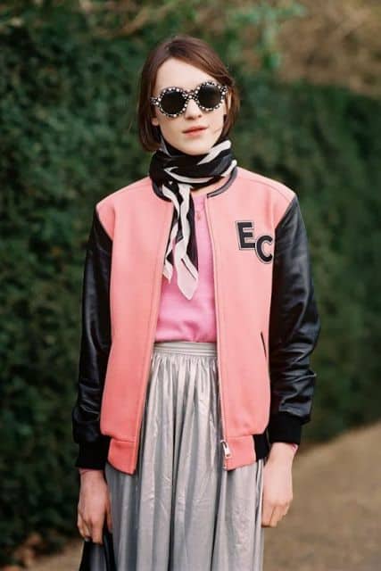 Modelo veste saia plissada, jaqueta rosa com manga de couro, óculos e blusa rosa.
