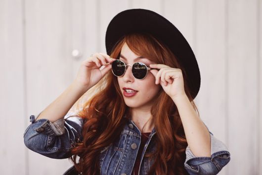 modelo usa óculos preto, chapeu na mesma cor e camisa jeans.