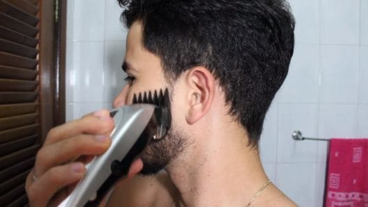 Homem de perfil passando a máquina de corte na costeleta, entre a barba e o cabelo