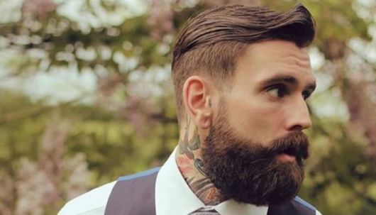 Homem de perfil com bastante tatuagens no pescoço e barba degradê estilo lenhador