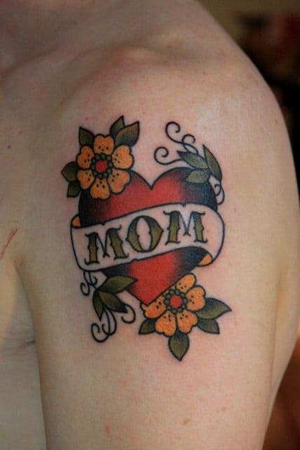 Tatuagem de coração no ombro. Ele é pintado de vermelho, possui flores ao seu redor e tem uma faixa no meio escrita "Mãe"