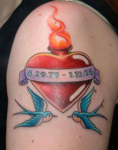 Tatuagem de um coração vermelho com chamas saindo dele e andorias voando a sua volta. No centro do coração há uma faixa com as datas "8.29.71 - 1.12.06"