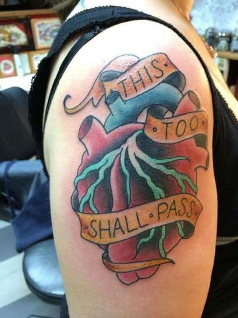 Tatuagem de um coração pintado de vermelho no ombro com uma faixa contendo os dizeres "Isso também passará" em inglês