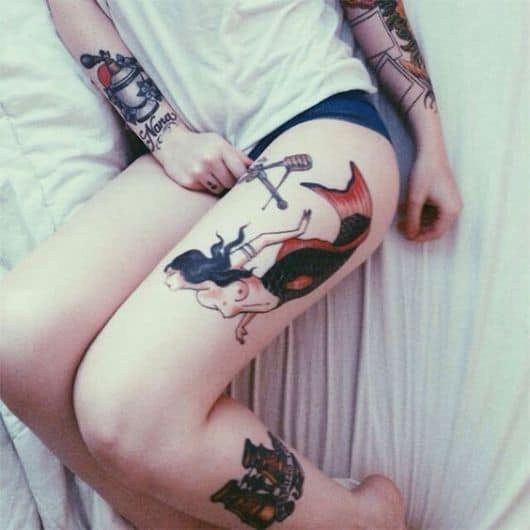 Tatuagem na perna de uma sereia nadando. Sua cauda é preta e vermelha e ela possui ornamentos no braço.
