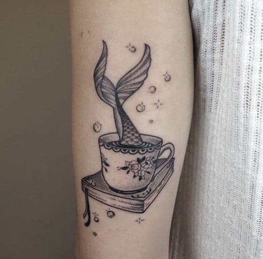 Cauda de sereia adentrando uma xícara de café apoiada em um livro