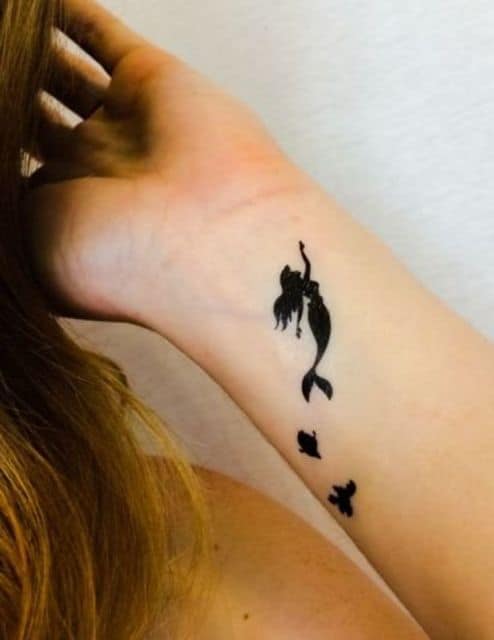 Tatuagem delicada no pulso da silhueta preenchida da princesa Ariel nadando acompanhada de seus amigos.
