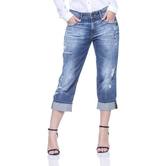 Modelo usa calça jeans azul com detalhes claros, camisa branca e sandalia preta.