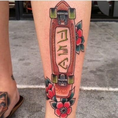 Tatuagem na canela de um longboard colorido apontando para cima e flores ao seu redor