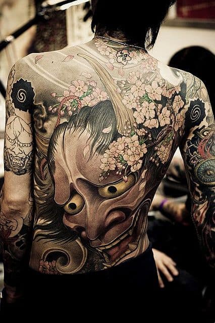 Tatuagem cobrindo completamente as costas de um homem de uma máscara hannya e diversas flores ao seu redor