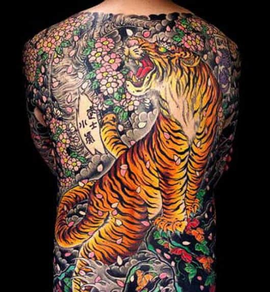 Tatuagem nas costas de um tigre rugindo e muitas flores e pétalas rosas em sua volta.