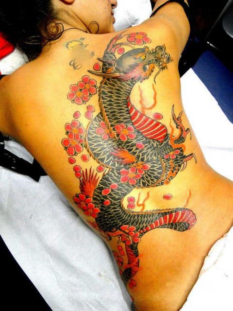 Tatuagem de um dragão oriental rodeado por flores feito nas costas de uma mulher que está deitada 