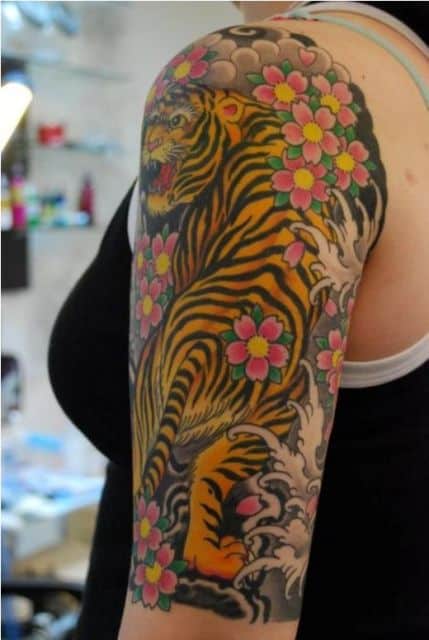 Tatuagem colorida de uma tigre visto de costas enquanto olha para trás feita na parte superior do braço de uma mulher
