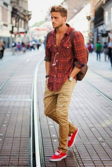 Homem caminhando na rua com as mãos no bolso vestindo uma camisa xadrez vermelha com as mangas dobradas e os botões superiores desabotoados 