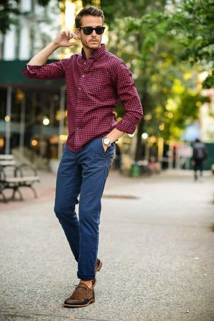 Homem na rua usando um óculos escuros, uma camisa xadrez vermelha com a manga dobrada, uma calça azul simples e relógio no pulso esquerdo 
