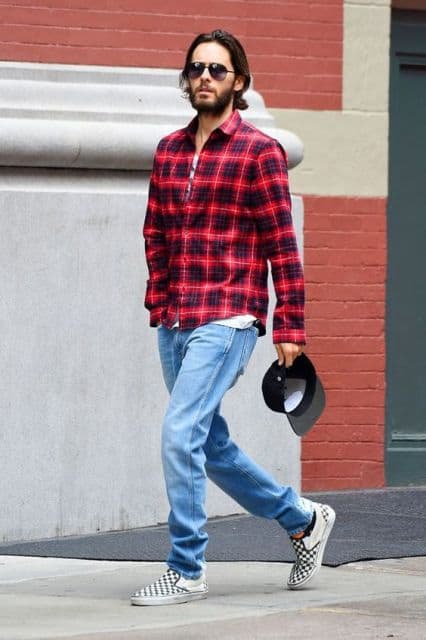 O ator Jared Leto caminhando na rua com uma camisa xadrez vermelha fechada, óculos escuros e uma calça jeans simples 