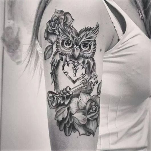 Tatuagem no braço de uma mulher de uma coruja com uma coração em seu peito e uma chave entre os dedos