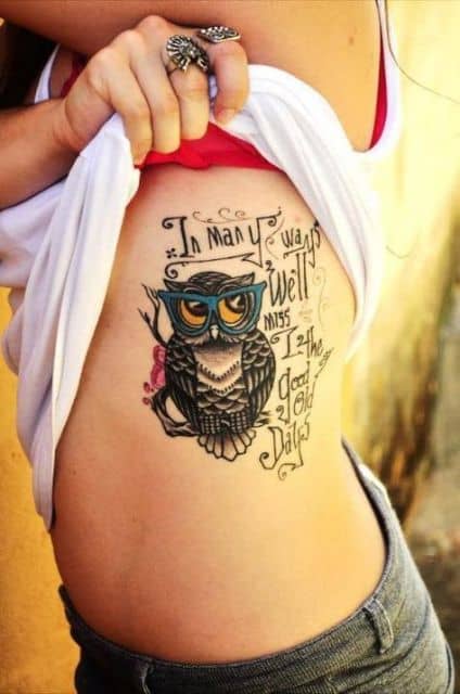 Tatuagem de uma coruja de óculos repousando em um galho com uma frase ao lado. 