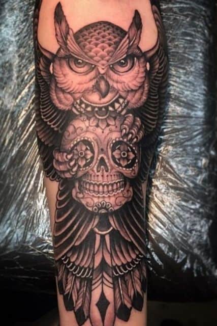 Tatuagem de uma coruja feita em preto e branco com as asas abertas segurando uma caveira mexicana entre suas garras.