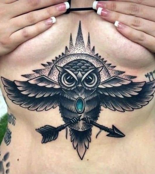 Tatuagem abaixo dos seios de uma mulher com uma coruja vista de frente enquanto voa com uma flecha quebradas em suas patas 