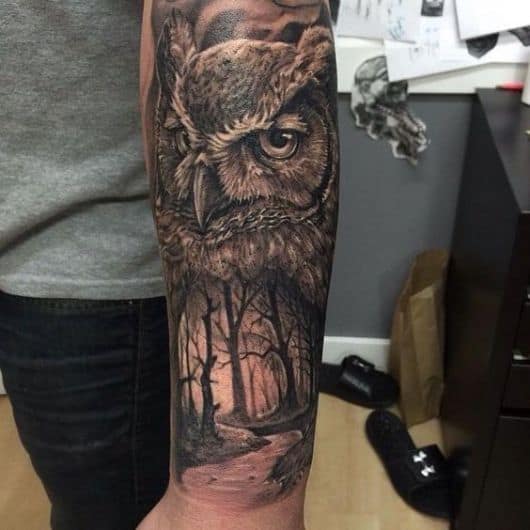 Tatuagem no antebraço de um homem com o rosto de uma coruja e uma floresta abaixo dela 