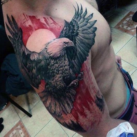 Tatuagem grande no braço de uma águia com asas abertas e a florestas e o sol atrás dela com tons de vermelho 