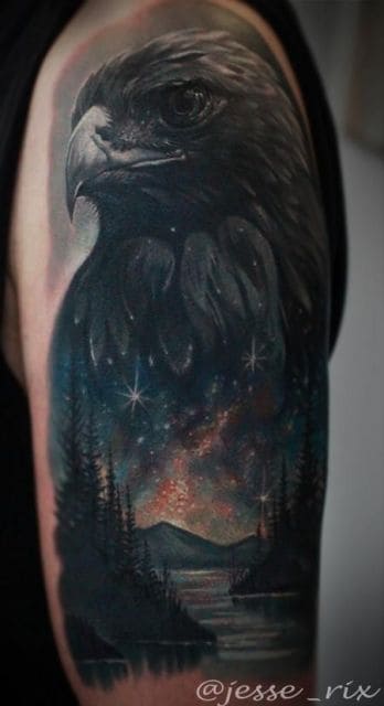 Tatuagem grande que vai do ombro até metade do braço de um homem com o desenho de uma águia e dentro de seu corpo há a ilustração de uma paisagem com montanhas e árvores 