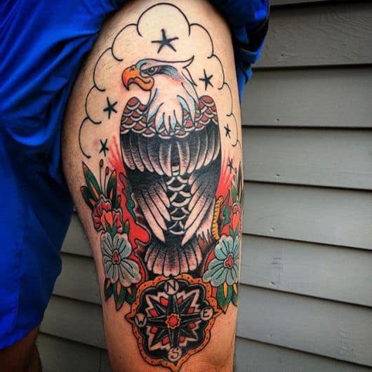 Tatuagem muito colorida de uma águia vista de costas com flores ao seu redor e uma bússola antiga abaixo dela