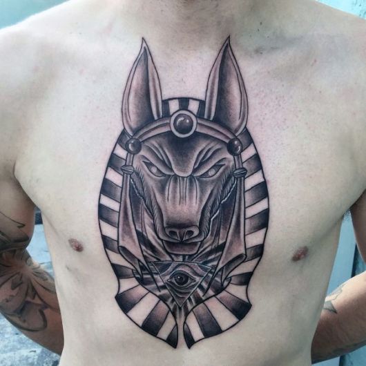 Tatuagem no peito de um homem do rosto do Deus Anúbis com a expressão brava feito em preto e branco.