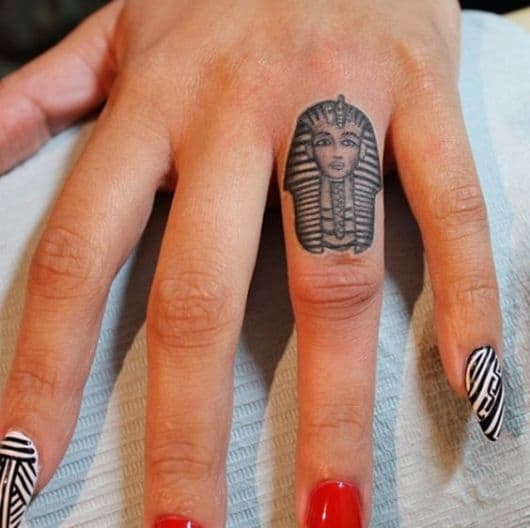 Tatuagem pequena do rosto de uma esfinge feita no dedo anelar. 