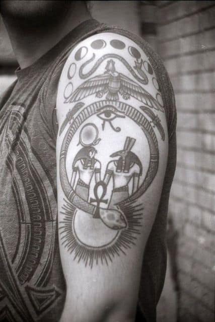 Tatuagem no braço de um homem com os deus Anúbis e Rá partilhando uma cruz Ankh envoltos por uma cobra. 