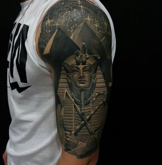 Tatuagem realista cobrindo a parte superior do braço de um homem com o desenho de uma esfinge e as pirâmides com um céu estrelado ao fundo. 