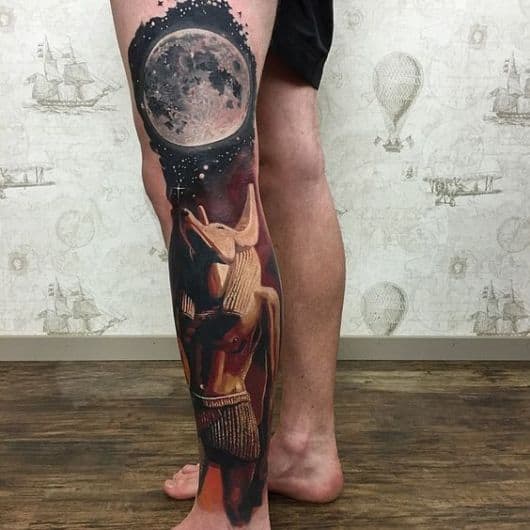 Tatuagem na perna de um homem com o desenho do deus Anúbis olhando para a lua. 