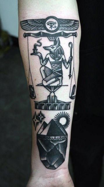 Tatuagem em preto e branco feita no antebraço com o desenho do deus Anúbis no centro de uma balança. 