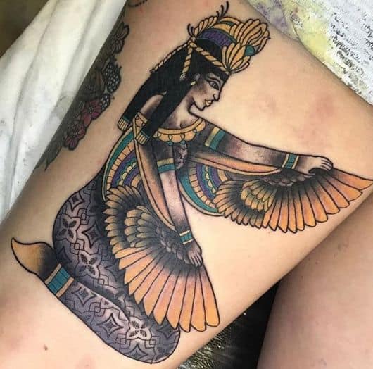 Tatuagem colorida feita na coxa com o desenho da Deusa Isís sentada segurando um tecido de penas em seus braços. 