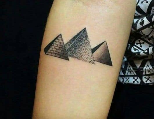 Tatuagem de três pirâmides, uma ao lado da outra. Uma tem as paredes lisas, outra tem as paredes com tijolos visíveis e a outra é formada a partir de diversos pontos conectados. 