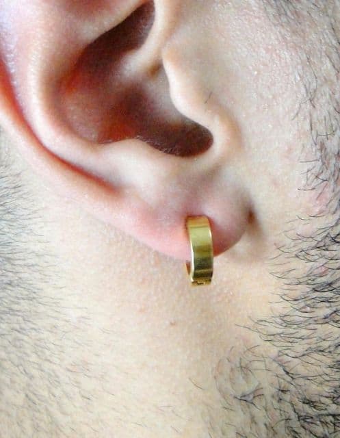 Foto em close up da orelha de um homem usando um brinco de pressão masculino dourado. 