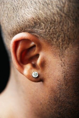 Foto em close up da orelha de um homem de pele negra e cabelo raspado com um brinco de pressão masculino brilhante. 