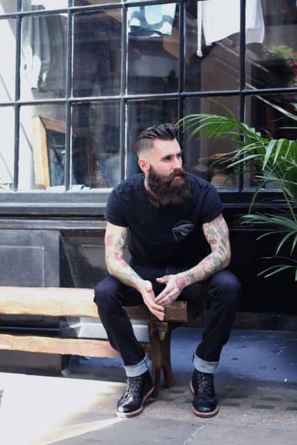 Homem tatuado sentado em um banco. Ele usa um undercut e barba muito grande. 