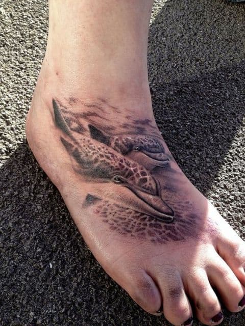 Tatuagem realista feita no pé com o desenho de dois golfinhos nadando lado a lado. 