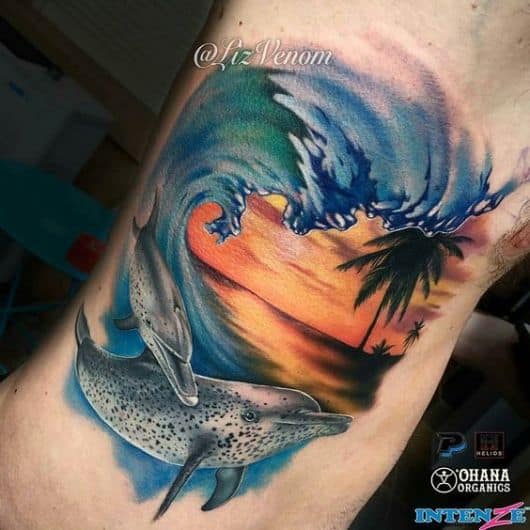Tatuagem de uma paisagem realista onde há dois golfinhos nadando em meio às ondas com o céu em estado de crepúsculo ao fundo. 