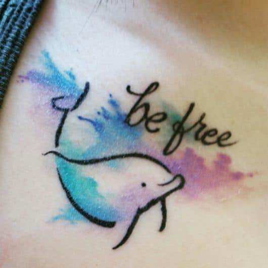 Tatuagem de golfinho feita no estilo aquarela com somente a silhueta do animal e a frase "Be Free'"