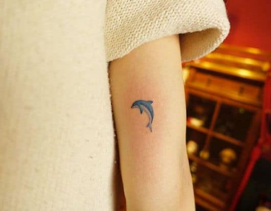 Tatuagem delicada de golfinho colorida com azul feita na parte de trás do braço