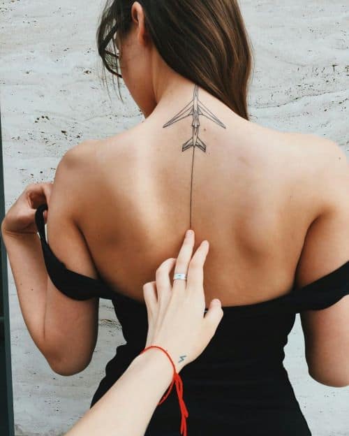 Tatuagem de um avião feito no centro superior das costas de uma mulher. Ele deixa um rastro que desce ao longo do centro de suas costas. 