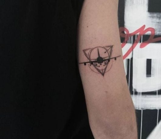 Tatuagem feia acima do cotovelo com a silhueta de um avião decolando dentro de um circulo que está inserido em um triângulo. 