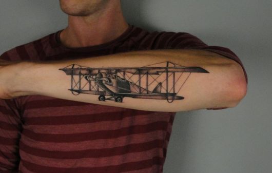 Tatuagem de um avião de modelo antigo feito na parte de fora do antebraço de um homem. 