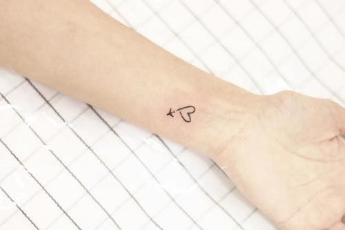 Tatuagem delicada no pulso de um avião voando e deixando um rastro que forma um coração. 