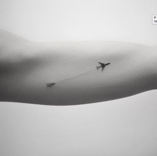 Tatuagem delicada do contorno de um avião preenchido de preto seguido por um avião de papel em sua traseira. 